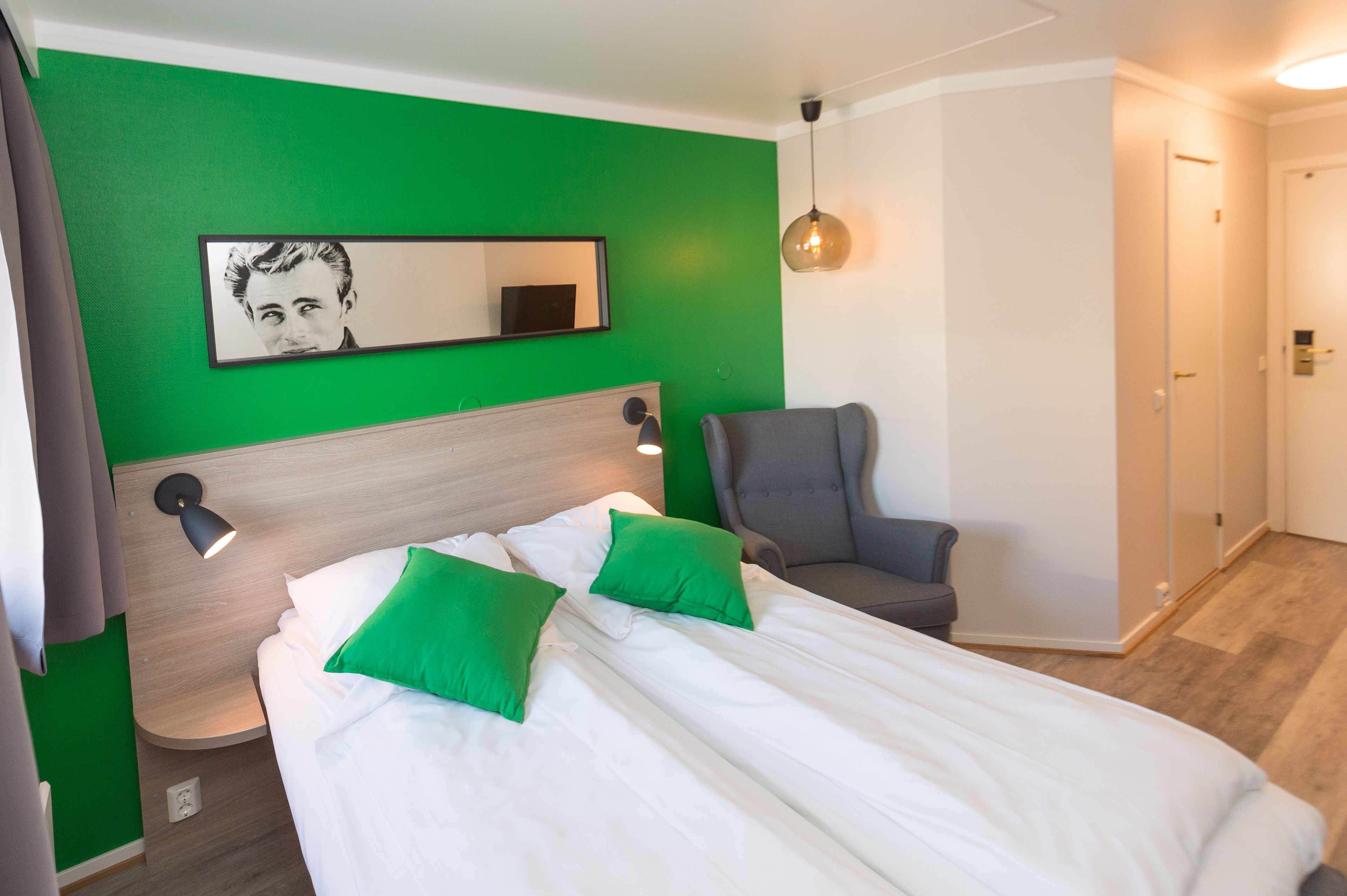 Ein Hotelzimmer mit zwei Betten, grünen Kissen, einem grauen Sessel, einem Spiegel und einer Pendelleuchte.