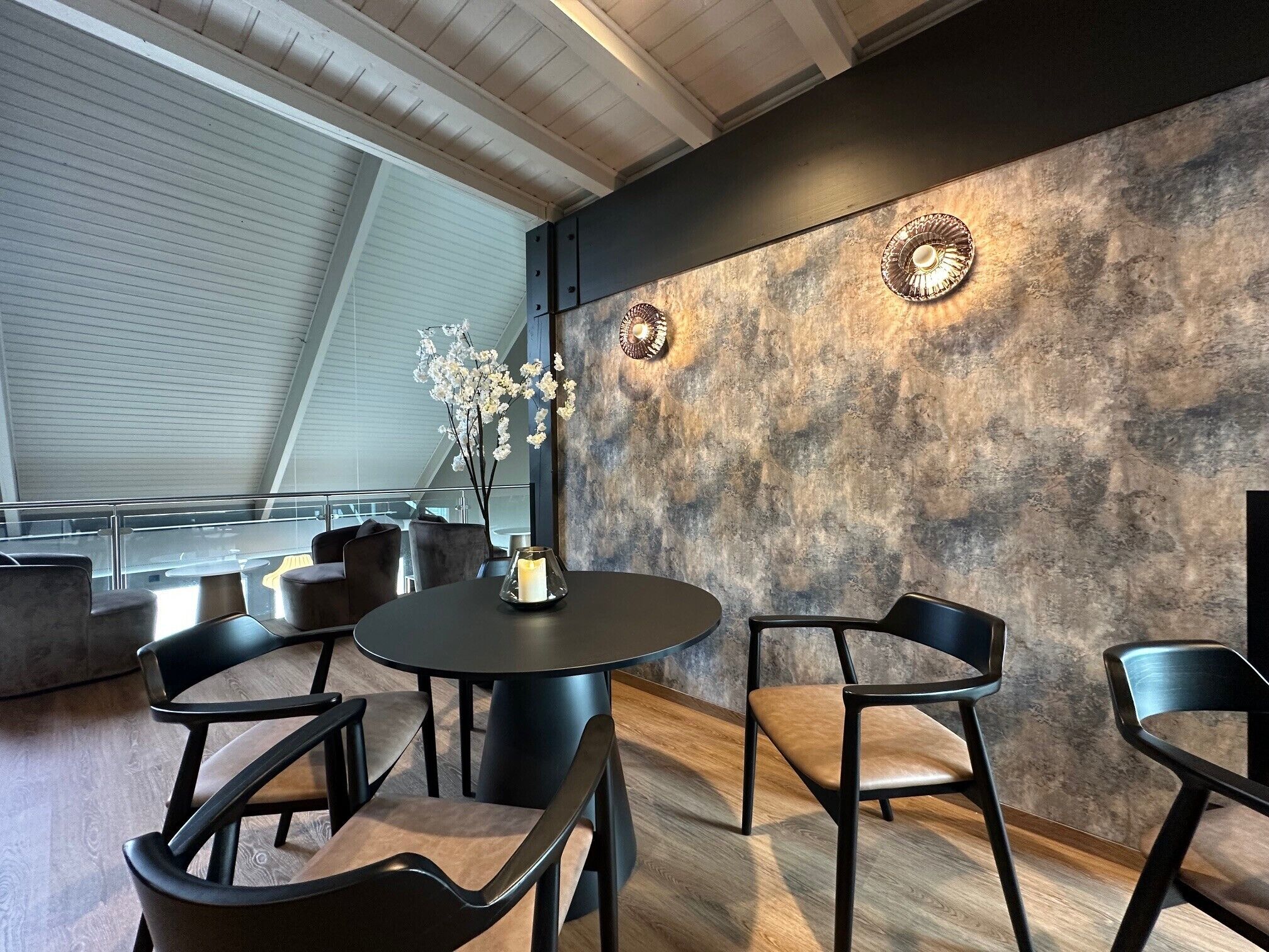 Een modern interieur met een ronde tafel, vier stoelen, een vaas met bloemen en een structuurwand met wandlampen.