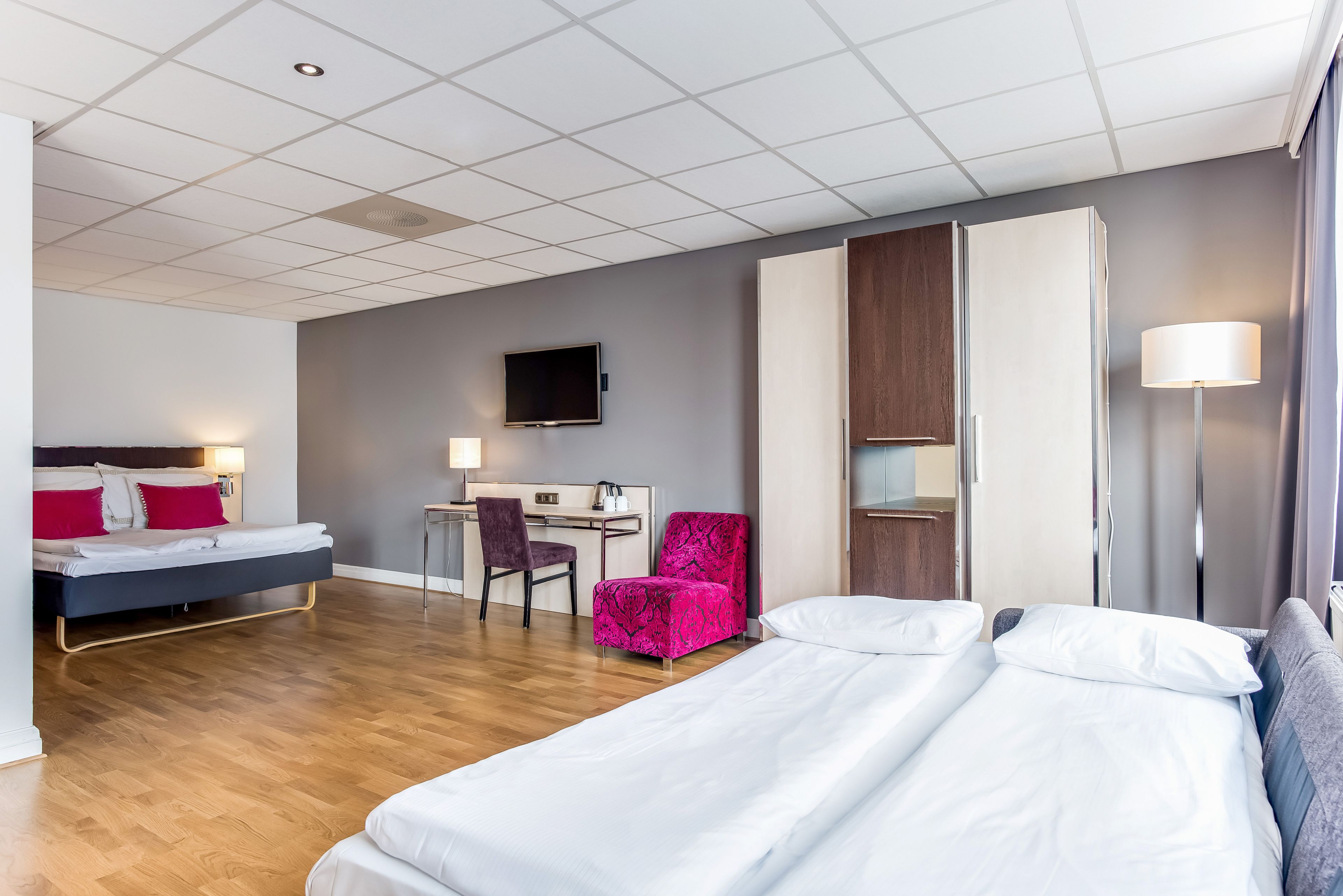A hotel room with a bed, desk, chair, TV, wardrobe, and lamp.