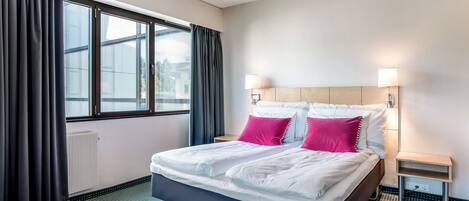 A hotel room with a large bed, two bedside tables, a window with curtains, and a radiator.