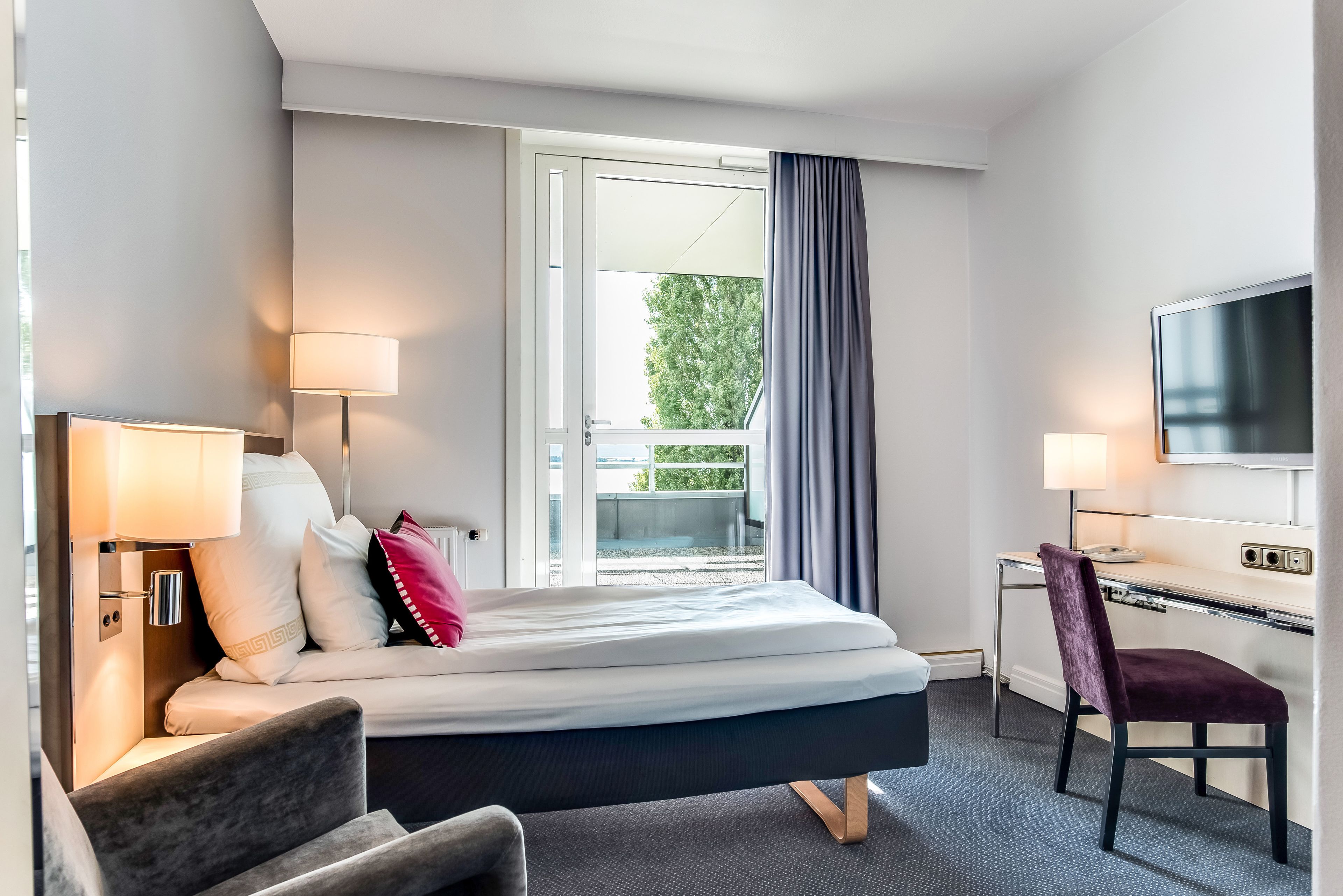 A hotel room with a bed, a desk, a chair, a TV, and a window with curtains.