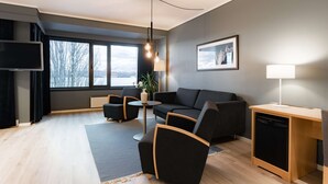 A modern living room with a sofa, two armchairs, a small table, a TV, and a window with a view of trees and mountains. - Scandic Alexandra Molde (Molde)