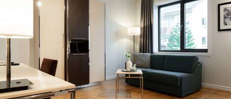 A modern living room with a sofa, a dining table with chairs, a small side table, a lamp, and a window with curtains.