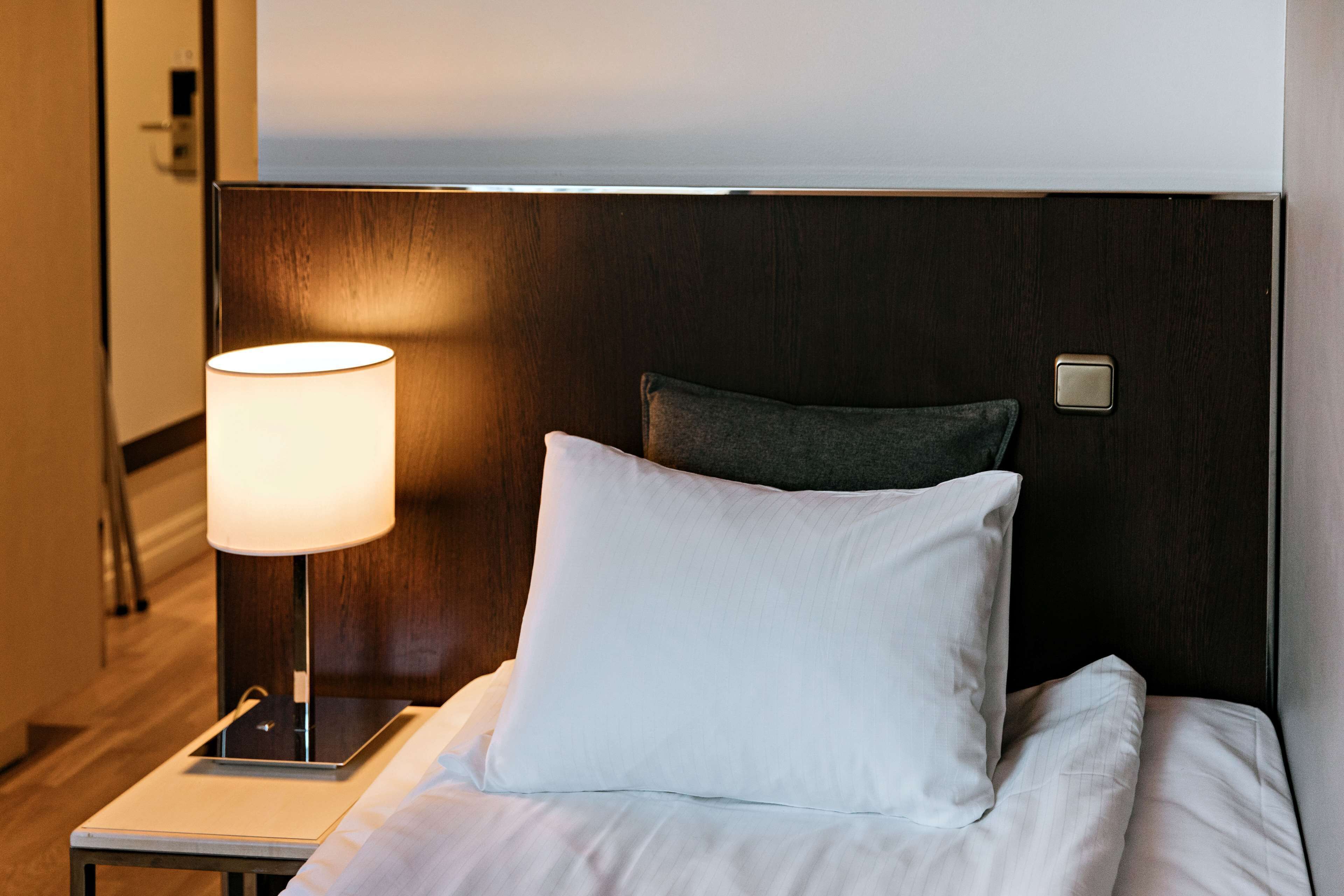 A neatly made bed with white linens and a single pillow, a bedside table with a lamp, and a wooden headboard.