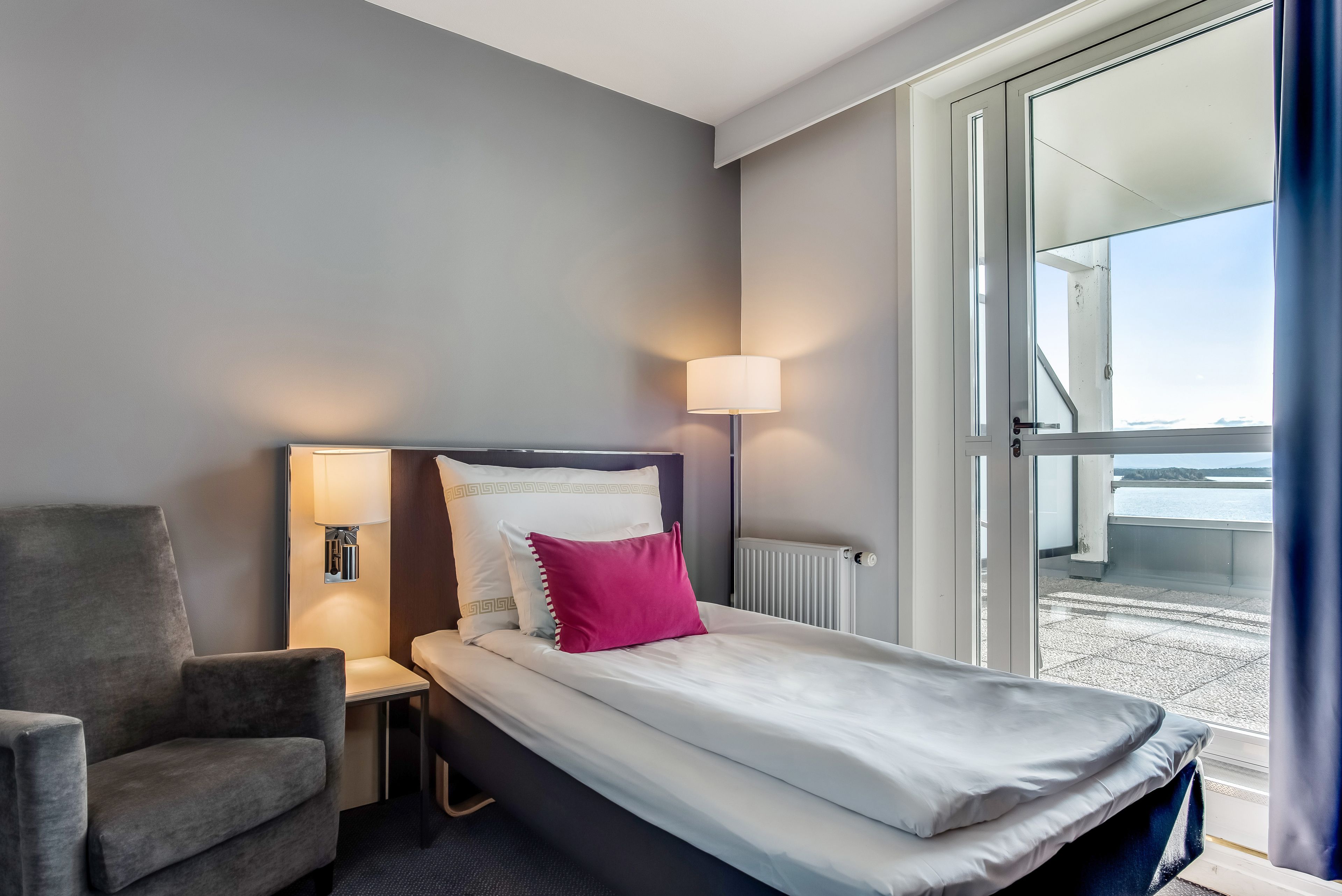 Habitación de hotel con cama, una silla, una mesita de noche, lámpara y vistas al exterior.