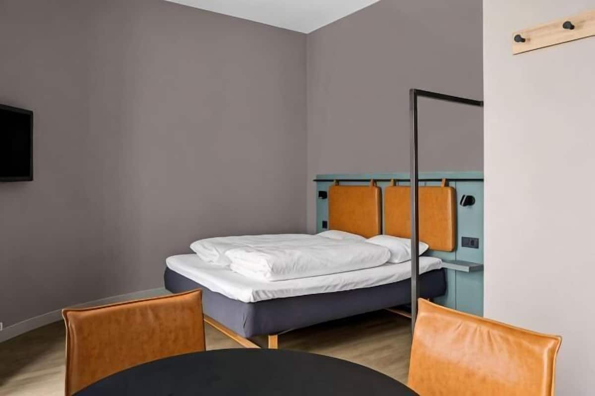 Una cama individual con cabecera, una mesa redonda y una silla en una habitación con pared gris y televisor montado.