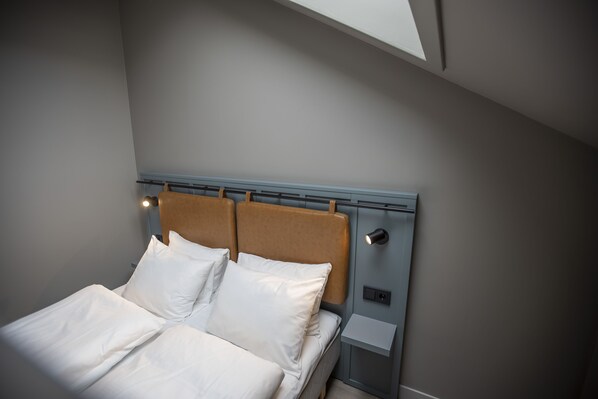 A neatly made bed with white linens and a brown headboard in a modern room with a skylight.