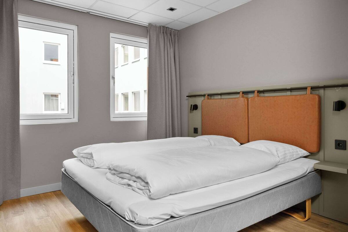 A neatly made bed in a room with two windows, a grey wall, and a wooden floor.