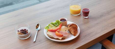 A breakfast setting with a croissant, a sandwich, a glass of orange juice, a glass of red juice, and a bowl of cereal on a wooden table.