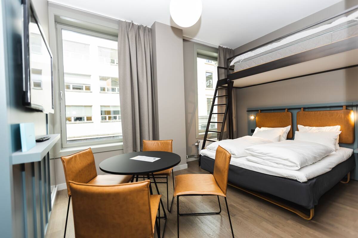 A hotel room with a bunk bed, a round table with chairs, a TV, and a window with curtains.