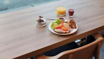 A wooden table with breakfast items including toast, a croissant, and a plate with vegetables and cheese. There are two glasses of juice and a small bowl of yogurt with fruit.