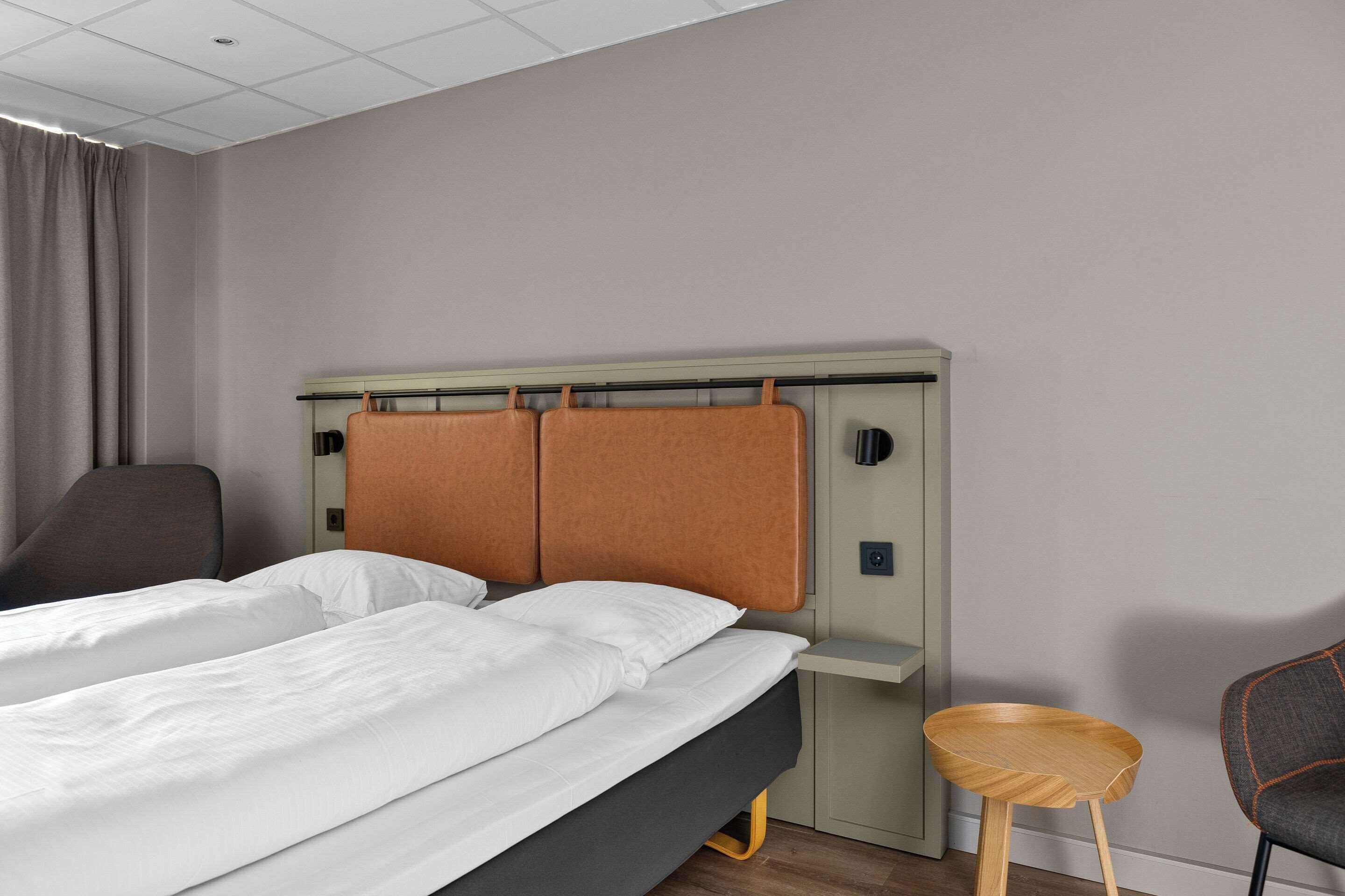 A hotel room with a bed, a desk, a chair, and a small stool.