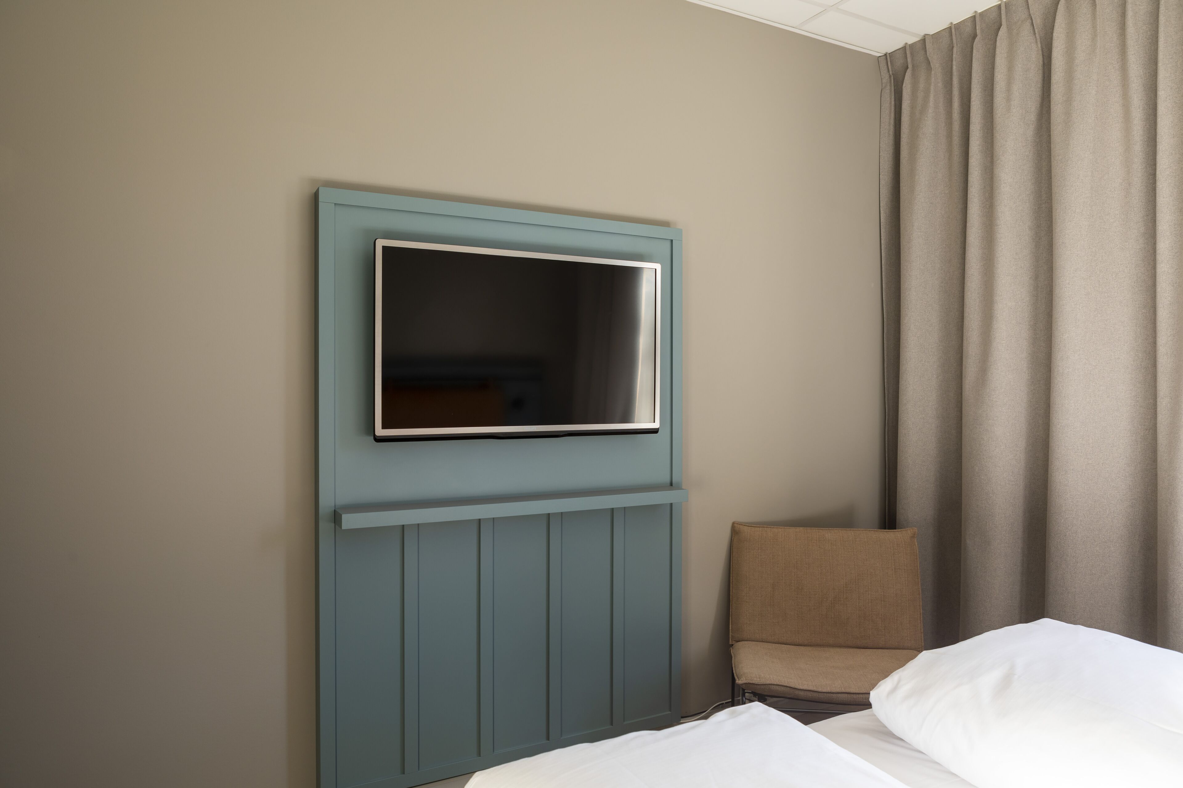 A hotel room with a bed, a chair, a television mounted on the wall, and a window with curtains.