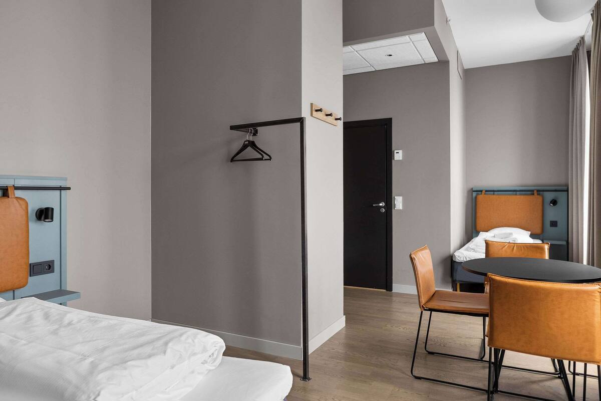 A hotel room with a bed, a desk with a chair, a dining table with chairs, and a door.