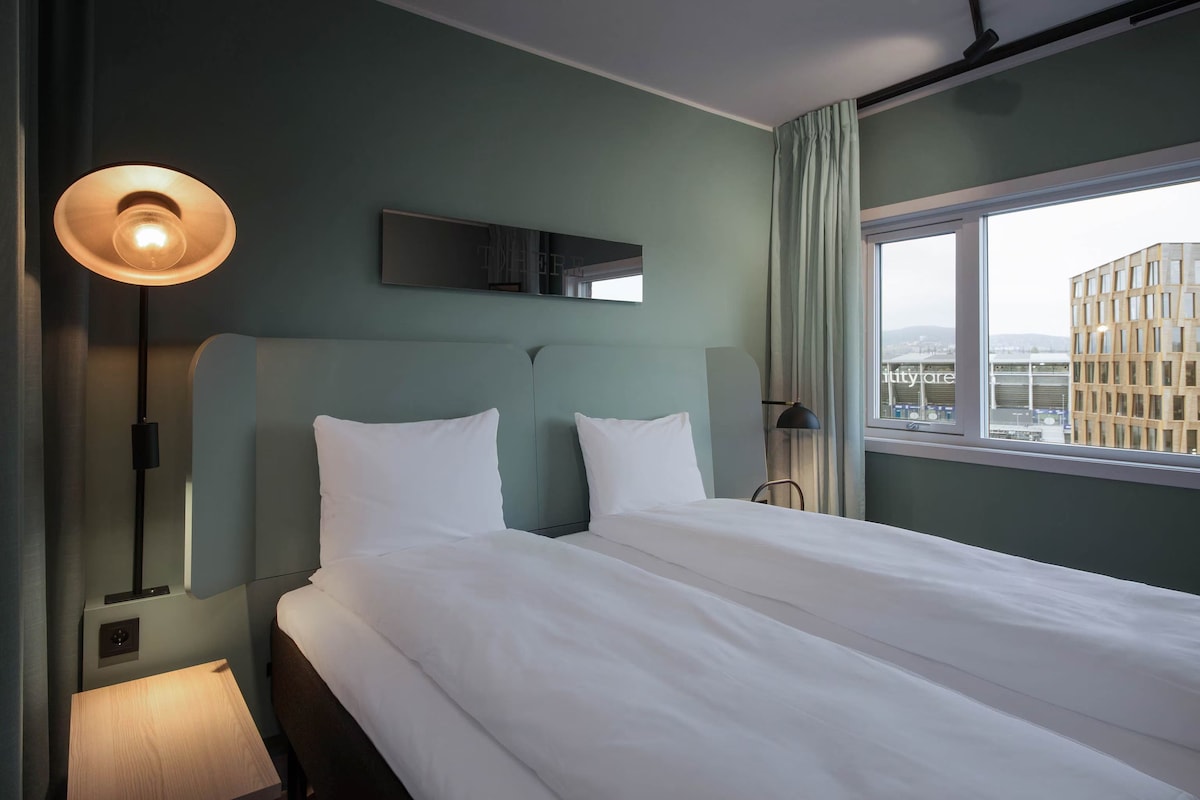 A hotel room with a large bed, a bedside table, a lamp, a mirror, and a window with a city view.