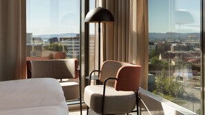 Una habitación de hotel con un ventanal grande, una cama, dos sillas y una lámpara de pie.