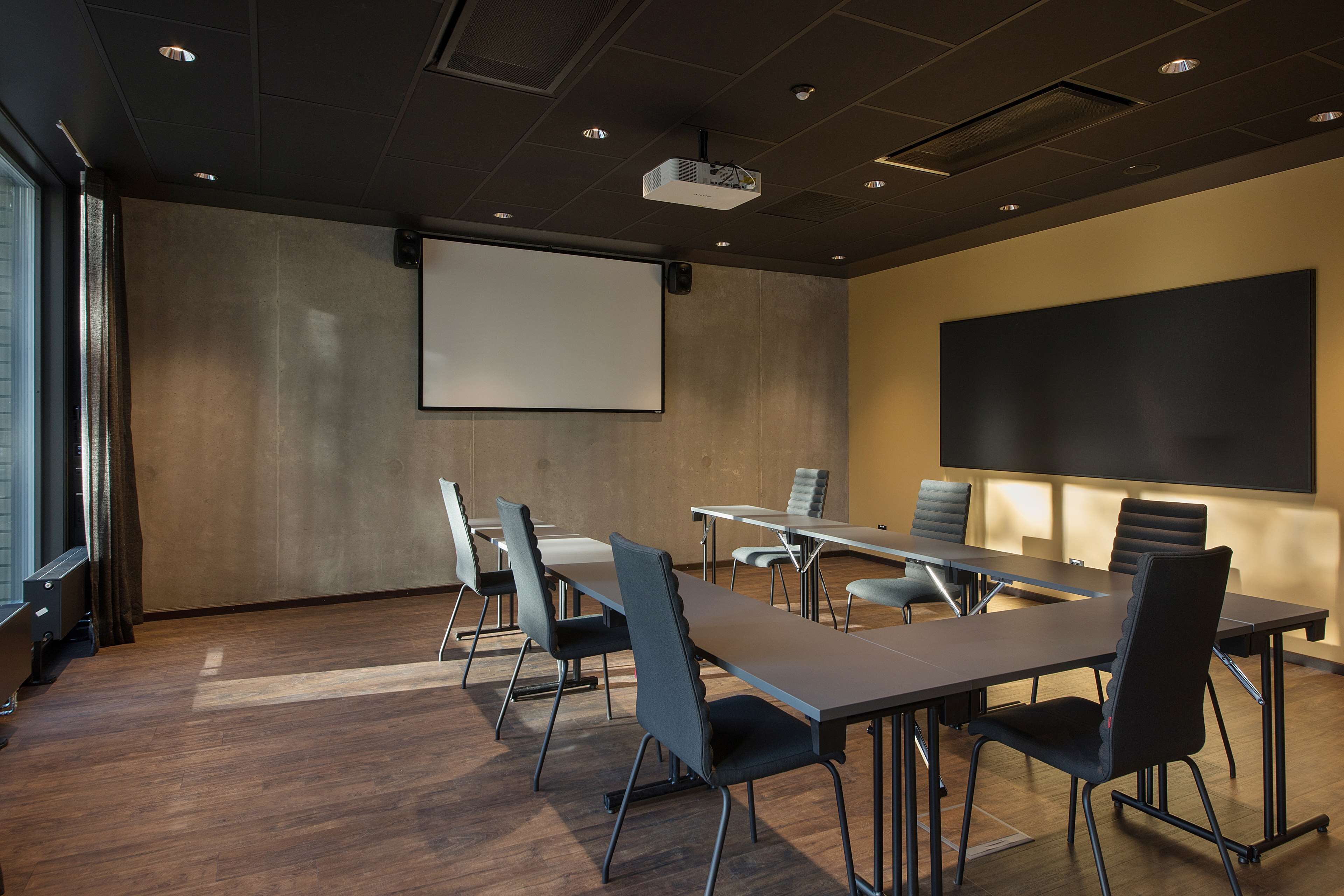 Una sala de conferències amb una taula llarga, diverses cadires, un pantalla de projector i una gran pantalla en blanc a la paret.