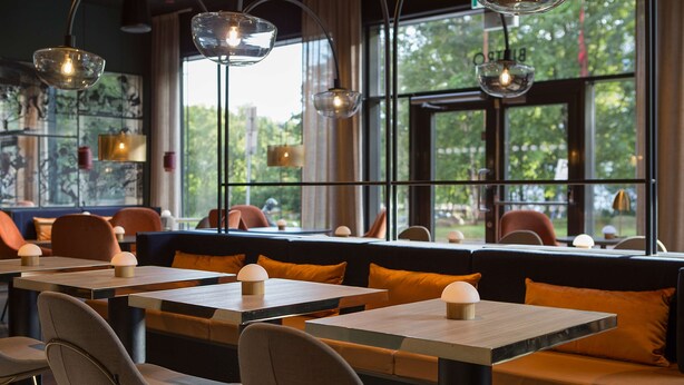 Interior de un restaurante con mesas, cabinas, iluminación colgante y amplios ventanales con vista a zonas verdes.