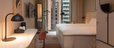 Una habitación de hotel moderna con una cama, mesita de noche y un escritorio con silla. Hay una ventana grande con vistas a un edificio.