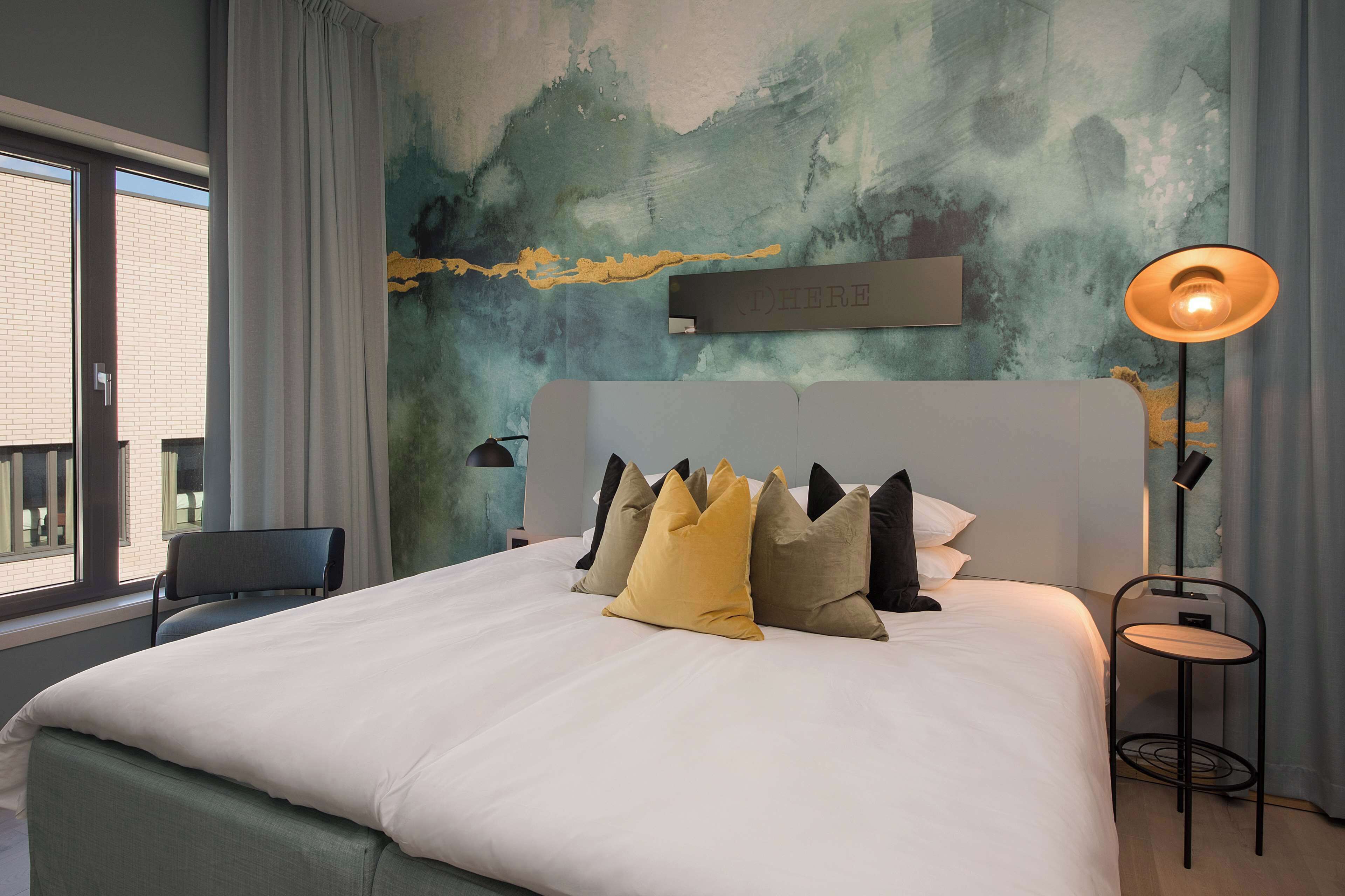 A neatly made bed with white bedding and multiple pillows, a modern lamp, and a wall with a textured abstract design.