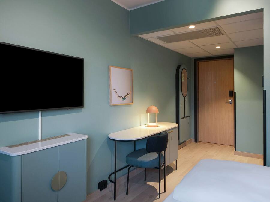 A hotel room with a bed, a desk with a chair, a television, and a door.