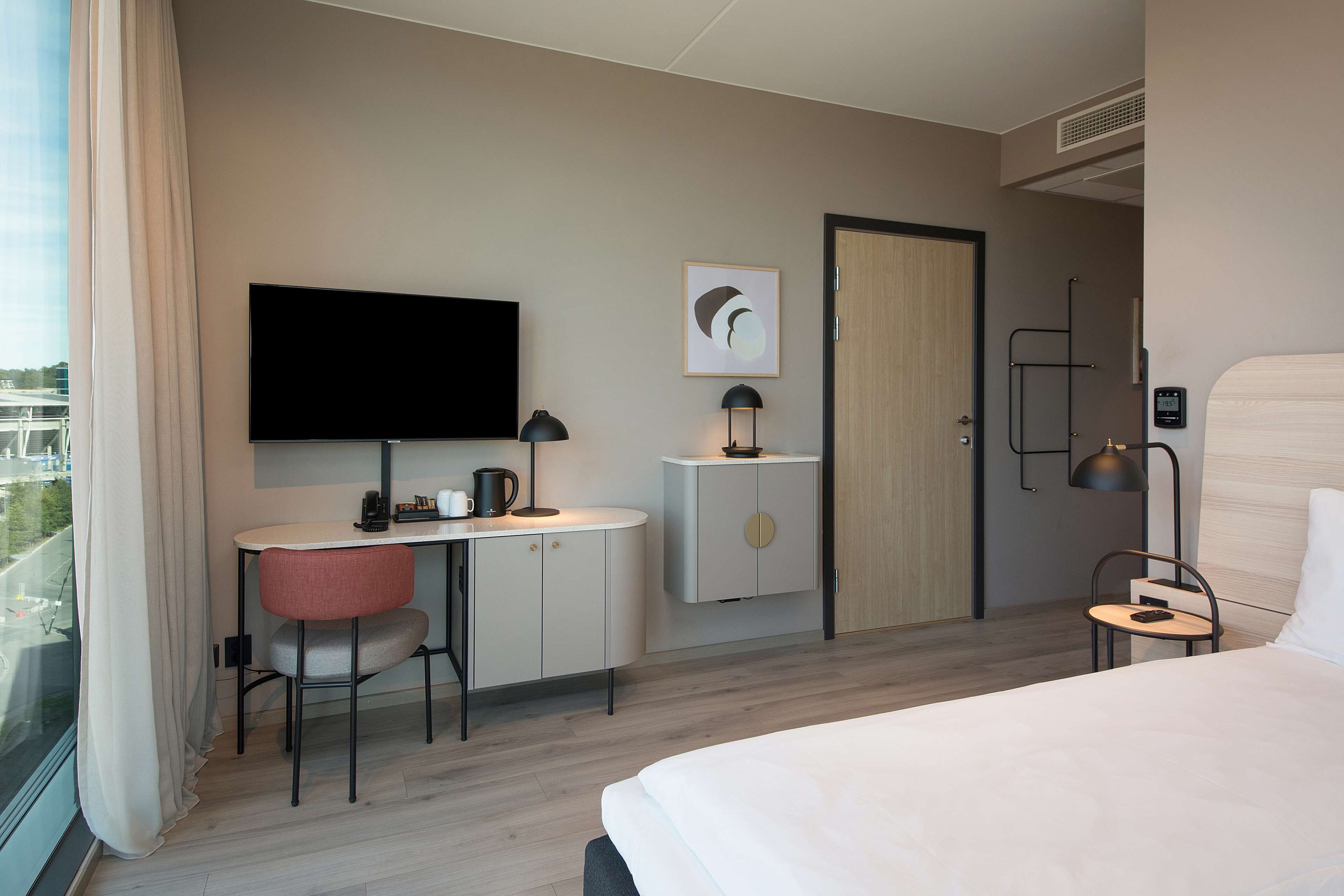 A hotel room with a bed, a desk with a chair, a television, a small table with a chair, and a door.