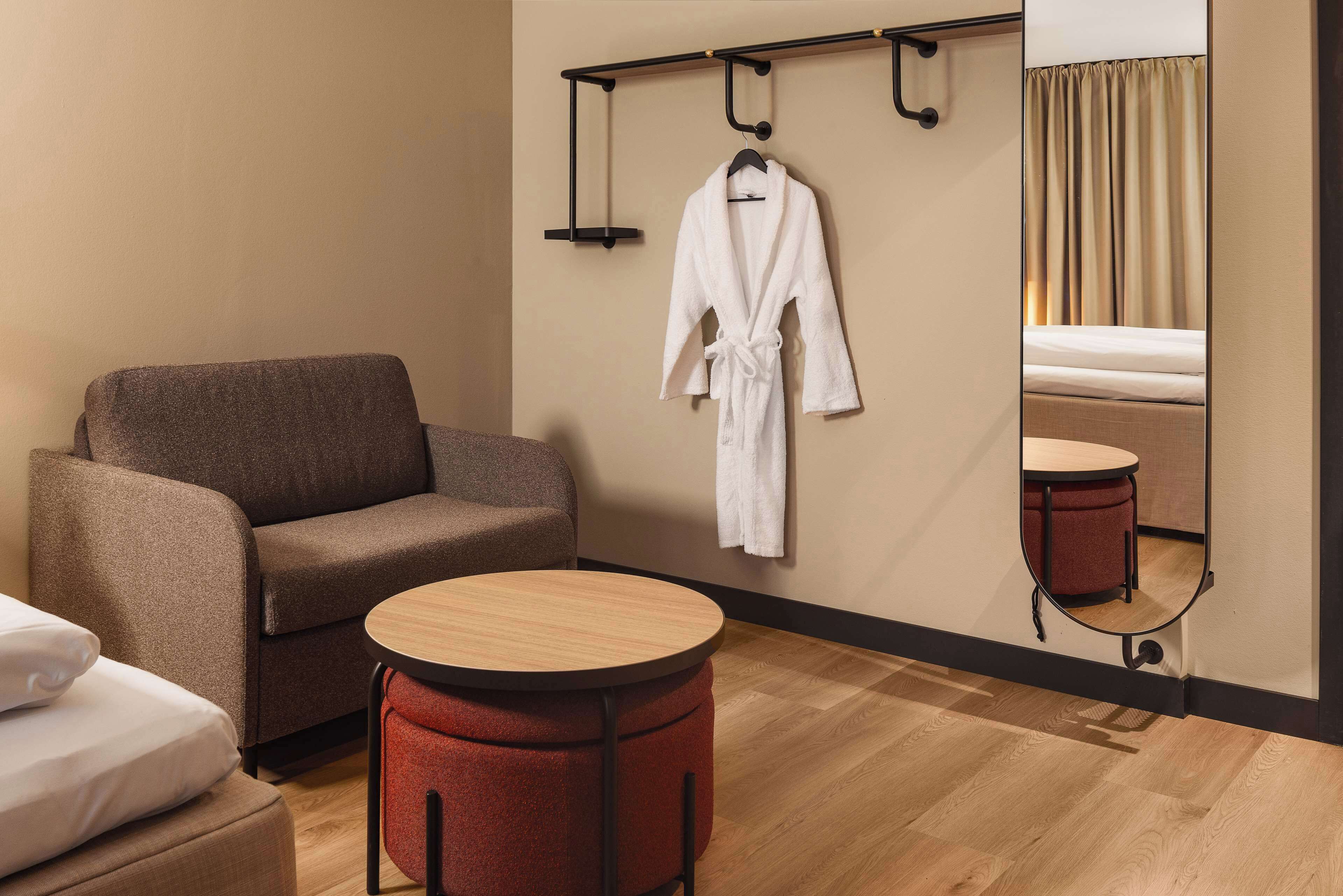 A hotel room with a sofa, a round table, a hanging robe, and a bed with a red ottoman.