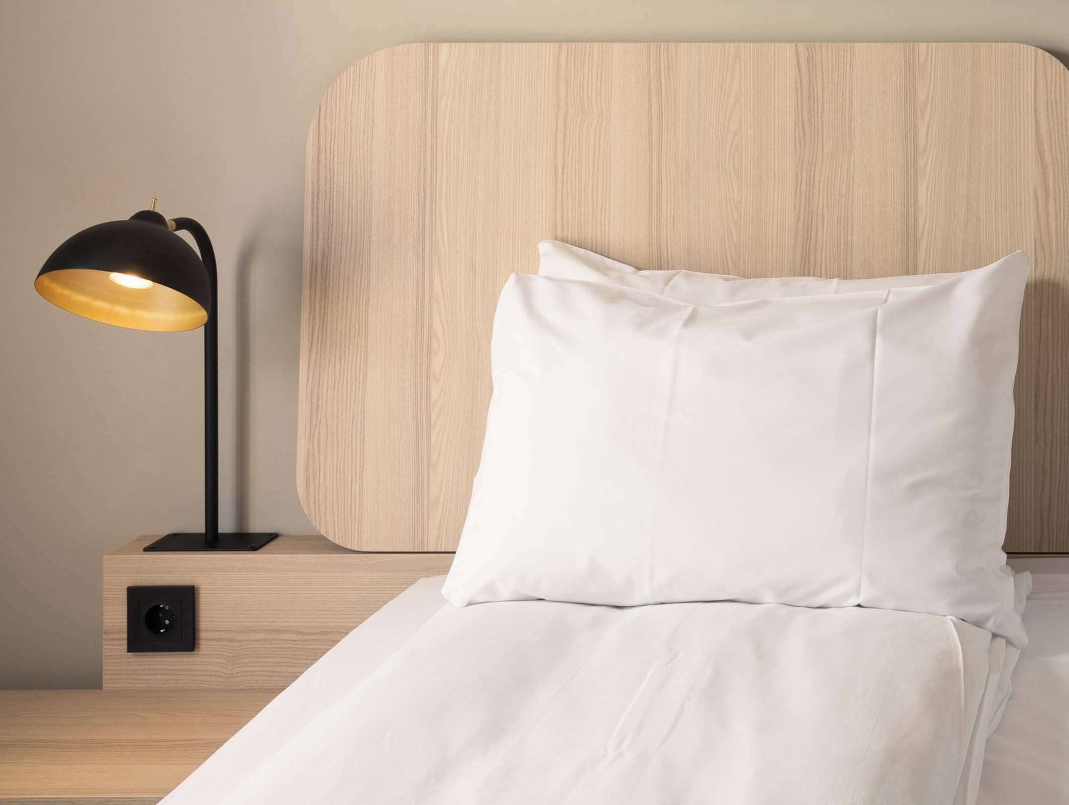 A neatly made bed with white bedding, a black floor lamp with a yellow shade, and a wooden headboard.
