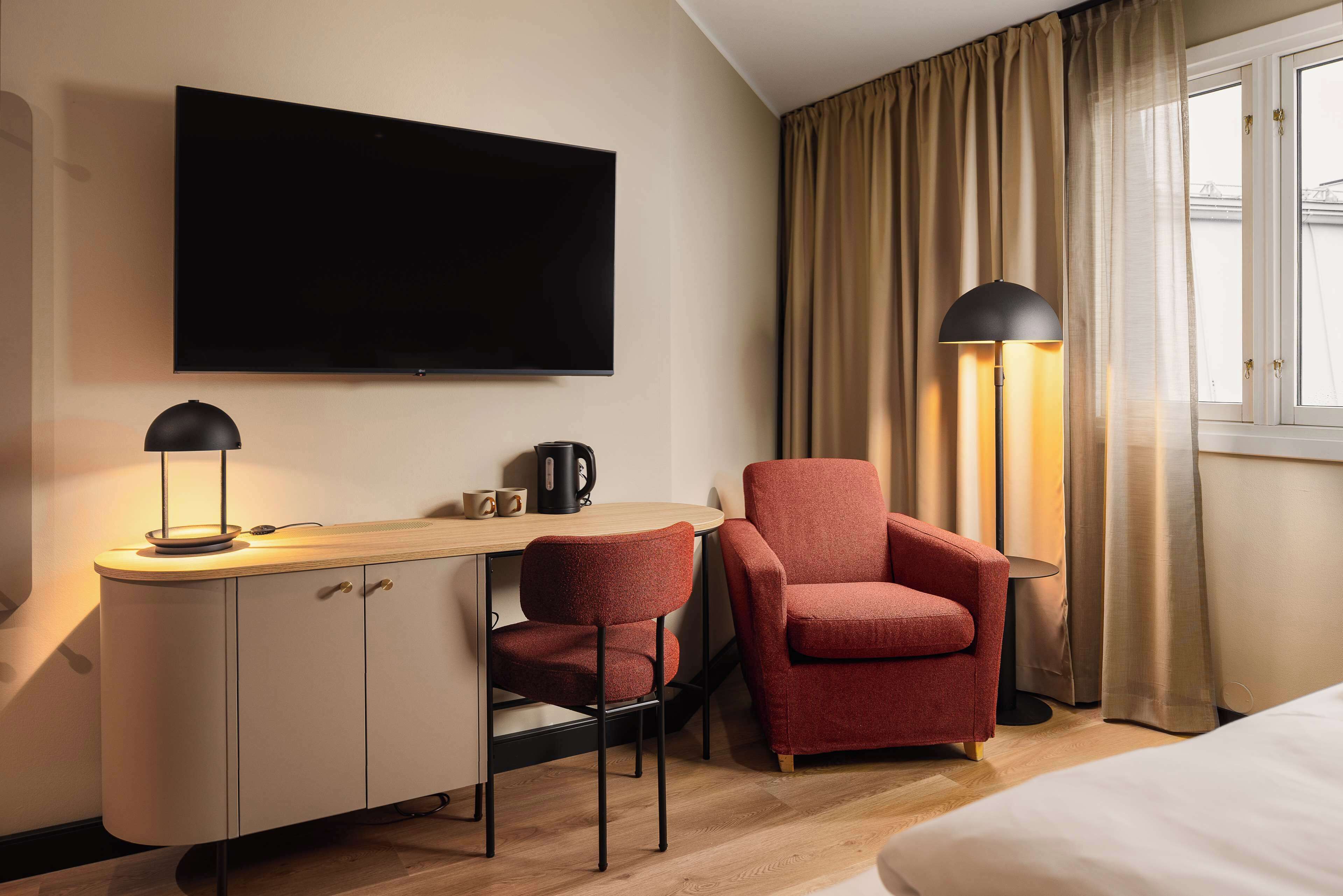 A hotel room with a flat-screen TV mounted on the wall, a desk with a lamp, a red armchair, and a bed.