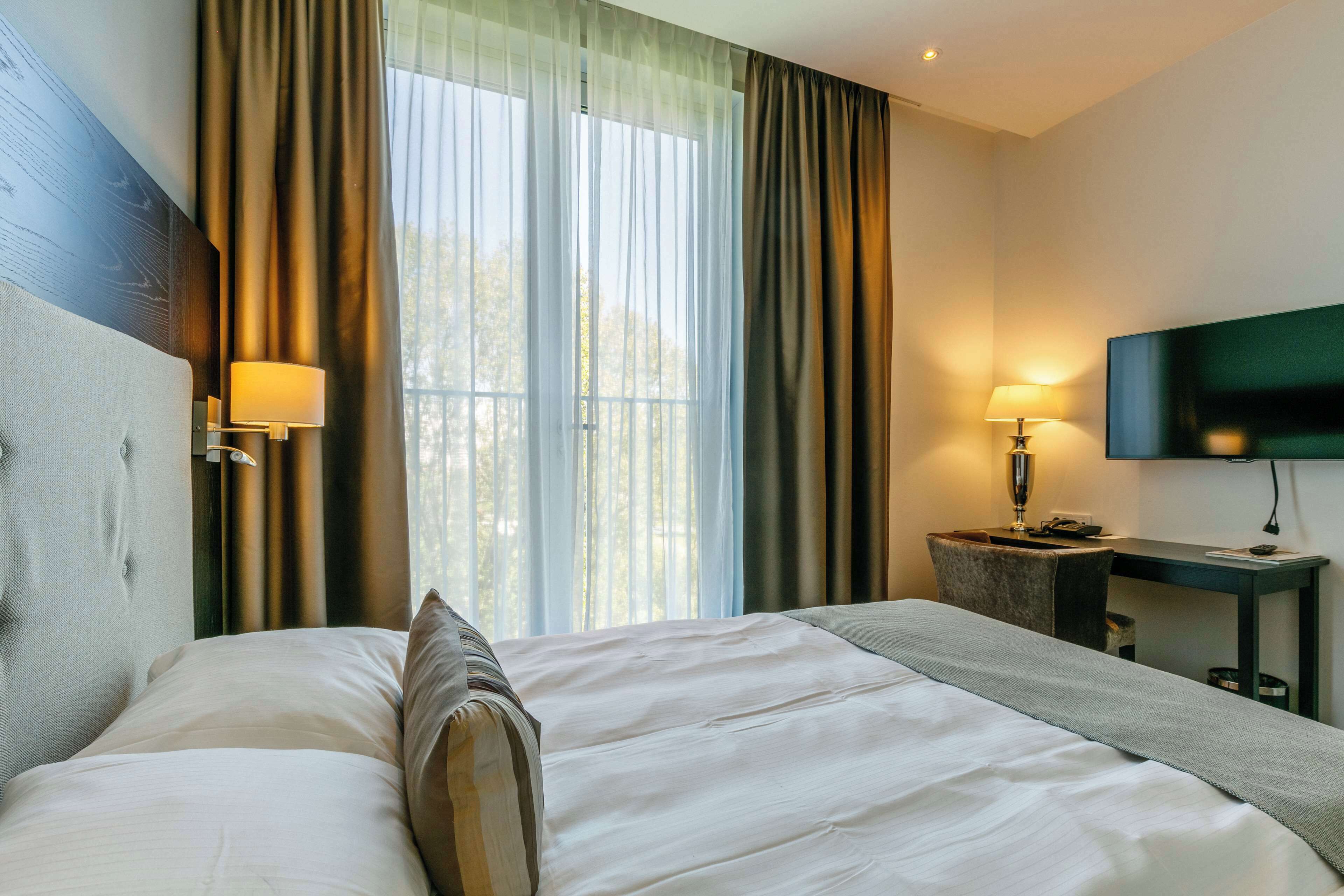 Ett hotellrum med en stor säng, ett skrivbord, en stol, en TV och gardiner.
