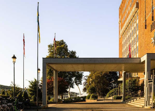 A covered entrance with a modern architectural design, featuring a brick building in the background and multiple flags on flagpoles. - Scandic Park Sandefjord (Sandefjord)