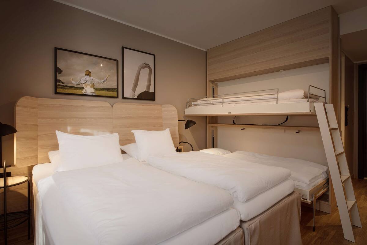 A hotel room with two beds, a bunk bed, a wooden headboard, and two framed pictures on the wall.