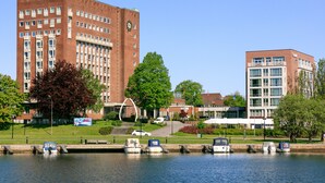 Vista del río con un campus que incluye un edificio grande de ladrillo, un edificio más pequeño adyacente y varias embarcaciones amarradas a lo largo del río.