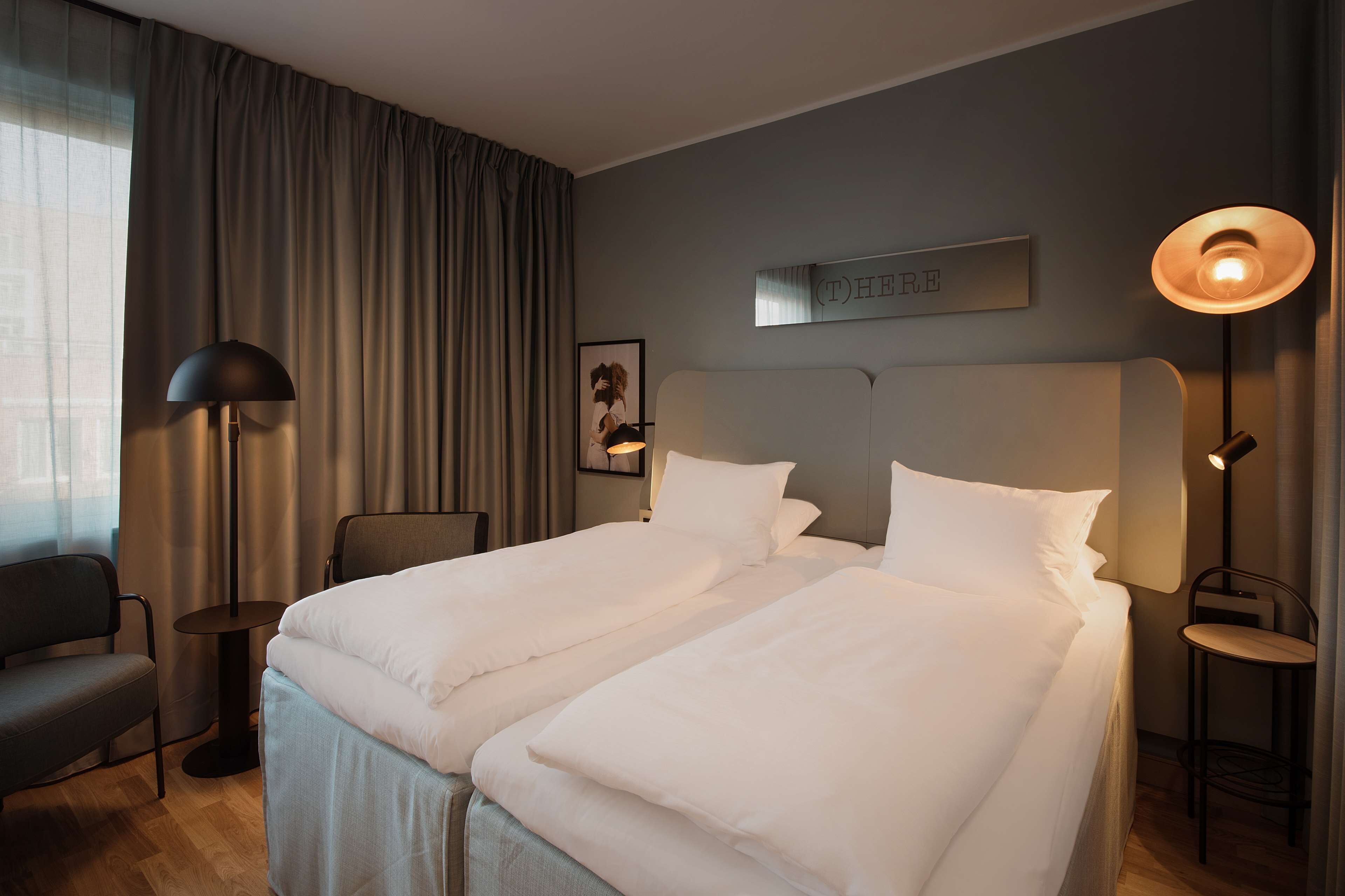 Ein Hotelzimmer mit zwei Betten, einem Schreibtisch, einem Stuhl, einer Lampe und einem gerahmten Bild an der Wand.