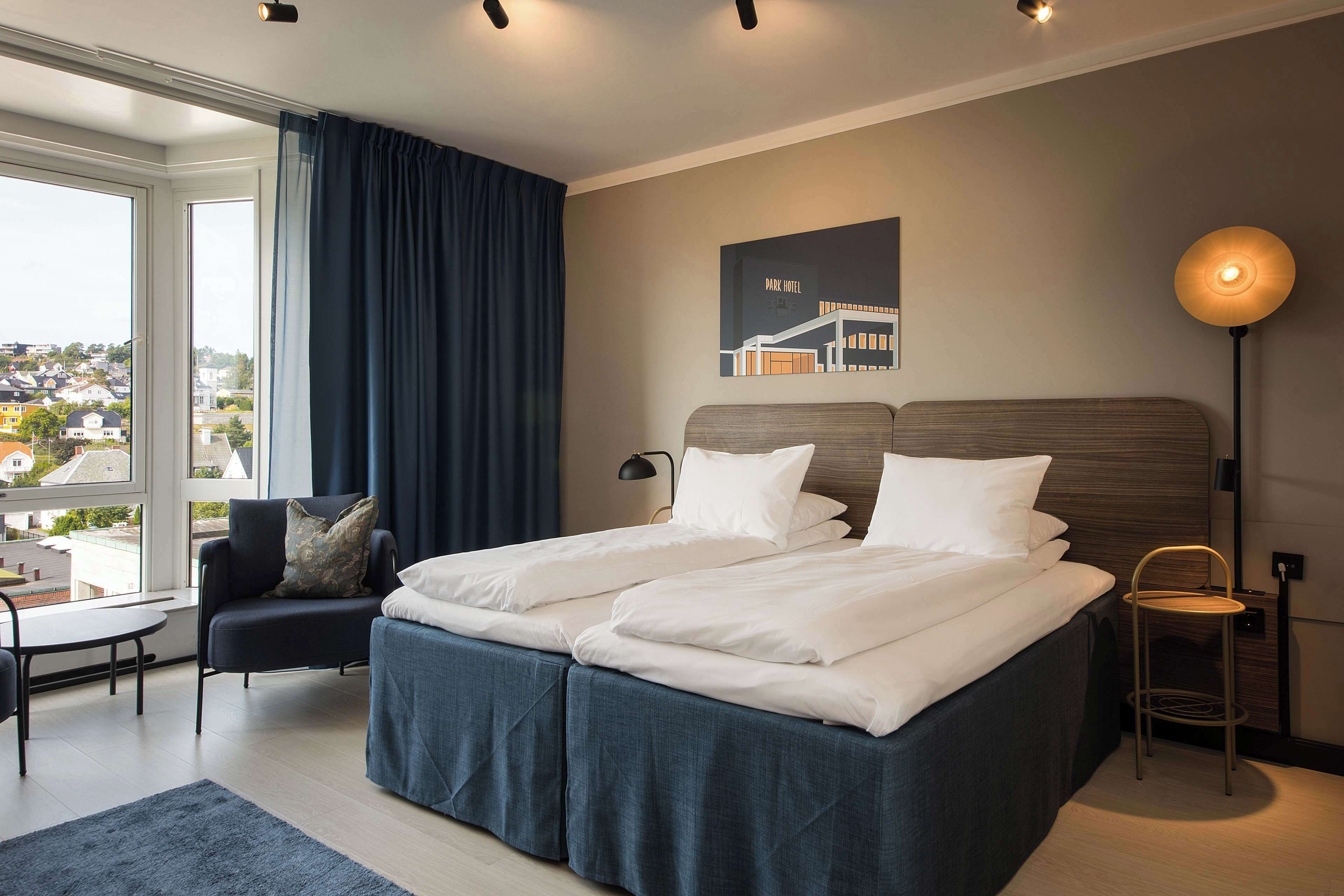 A double bed with white linens, a dark blue headboard, a small table, a chair, a floor lamp, and a cityscape view through a large window.