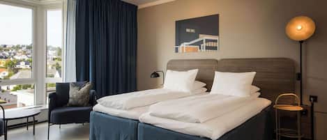Una cama doble con sábanas blancas, un cabecero azul oscuro, una mesita, una silla, una lámpara de pie y vistas a un paisaje urbano a través de un ventanal grande.