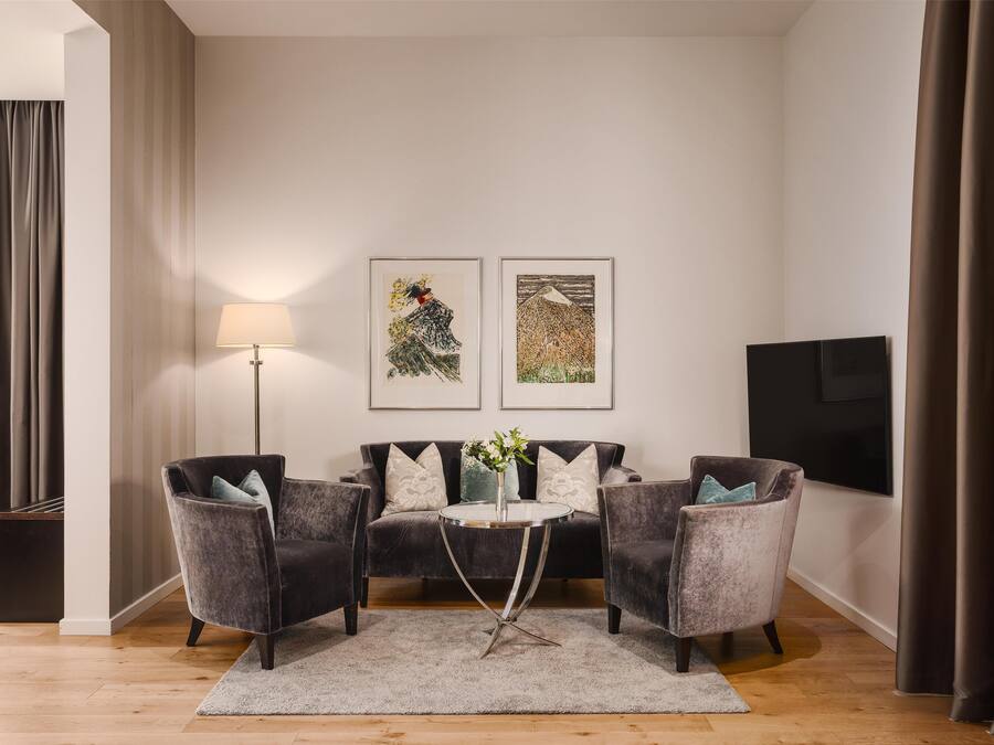 A modern living room with a dark grey sofa, two armchairs, a small round table, and a floor lamp. There are two framed artworks on the wall and a flat-screen TV to the right.