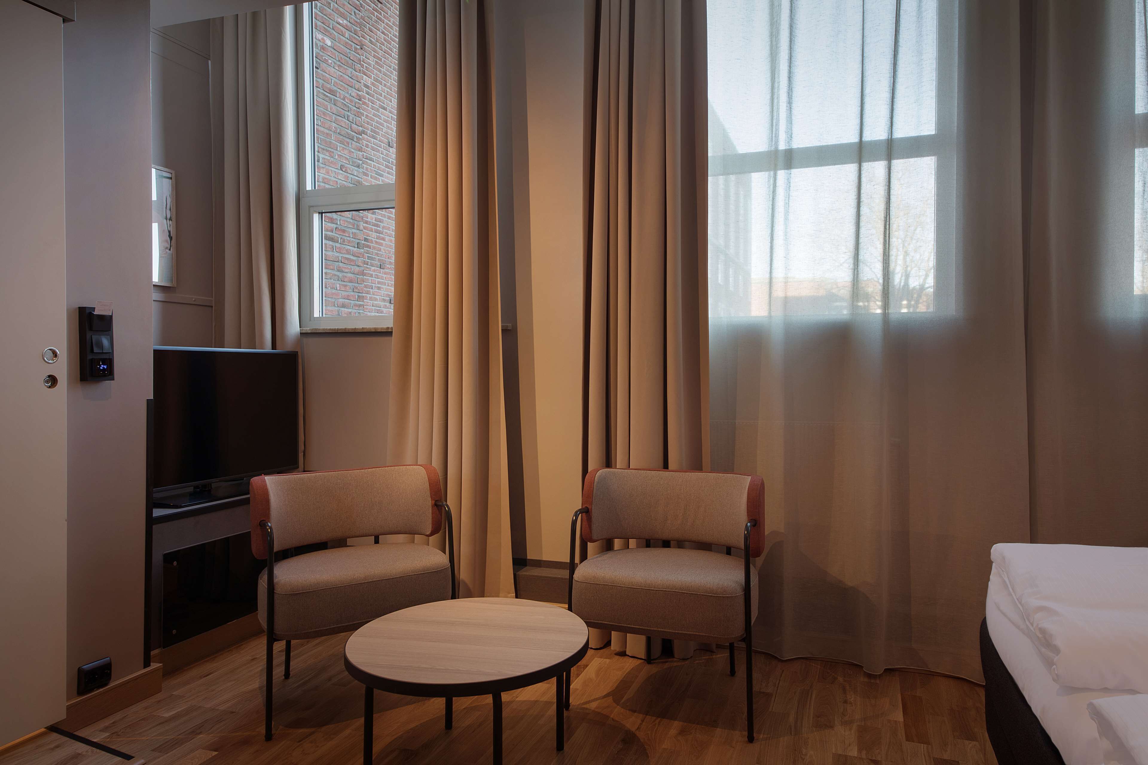 A hotel room with a wooden floor, two armchairs, a small round table, a bed, a TV, and a window with curtains.