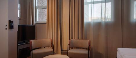A hotel room with a wooden floor, two armchairs, a small round table, a bed, a TV, and a window with curtains.
