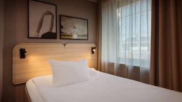 A hotel room with a wooden headboard, a white bed, two framed pictures on the wall, and a view through sheer curtains.