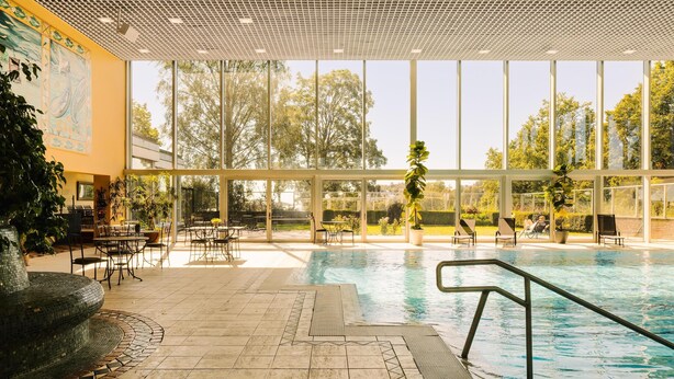 Una piscina interior con paredes de vidrio transparente, suelo de baldosas y zonas de estar al aire libre.