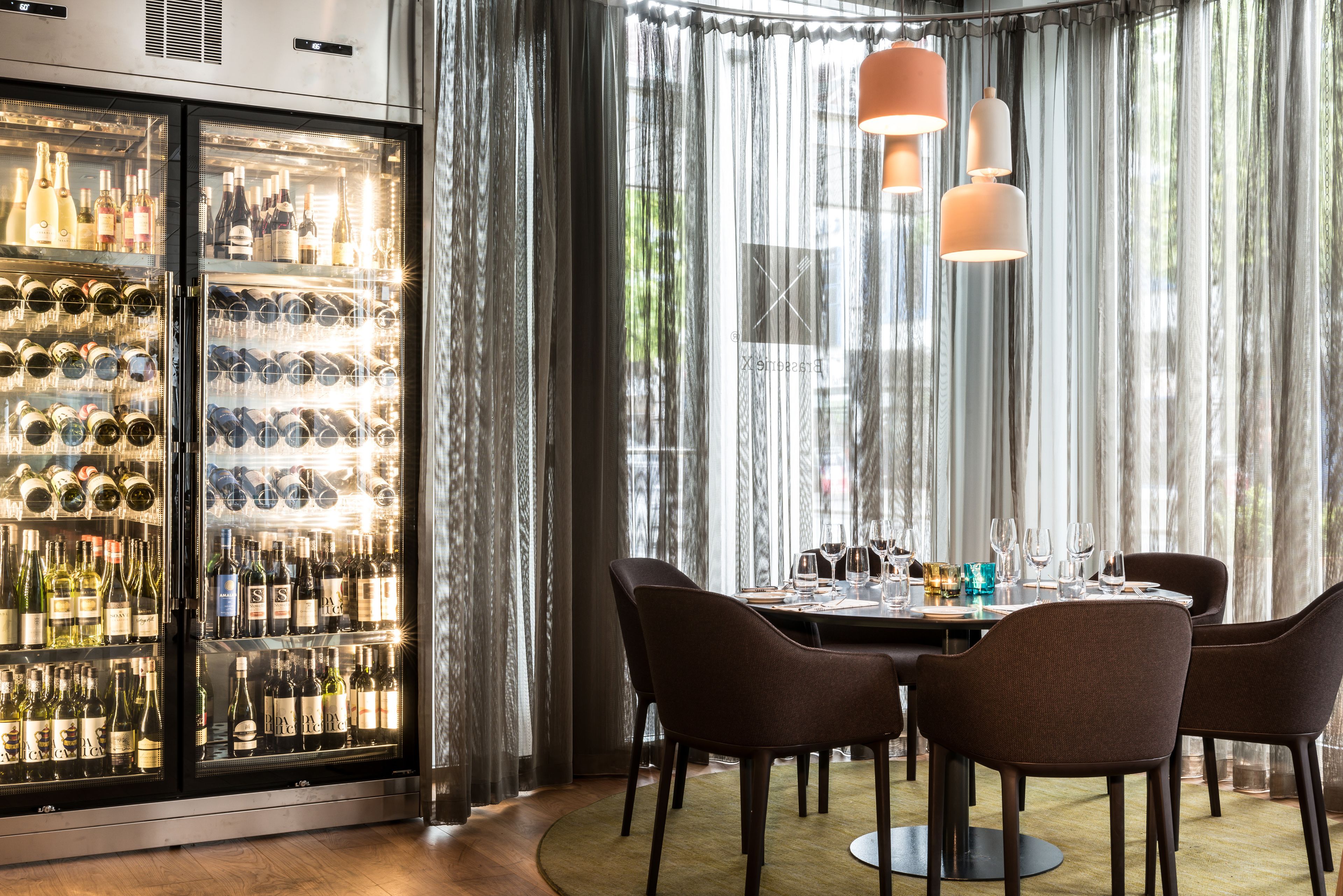 Restaurante com um refrigerador de vinhos, mesa posta para refeição e vista através de cortinas translúcidas.