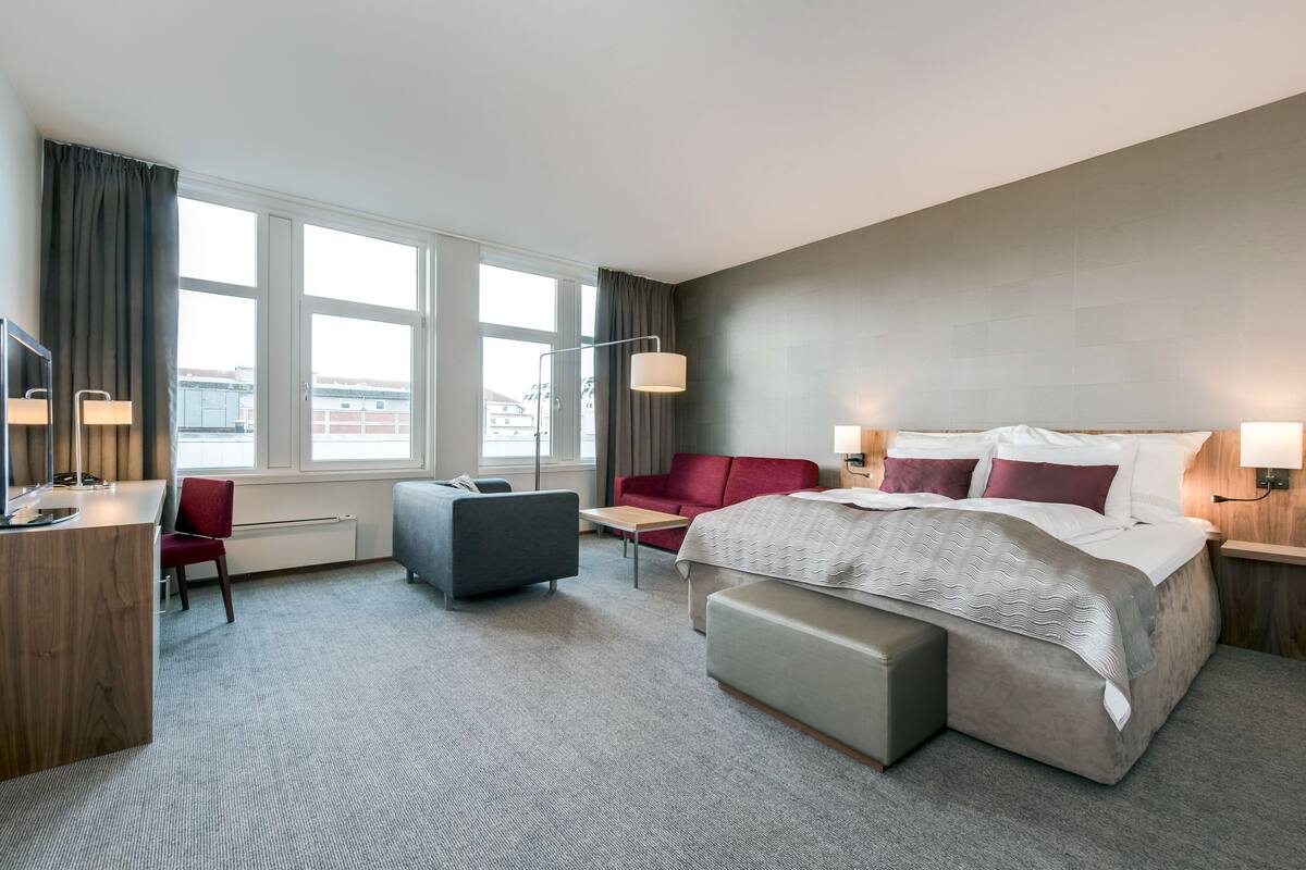 A modern hotel room with a large bed, a desk, a chair, and a sofa. There are two windows with curtains, a lamp, and a view of buildings outside.