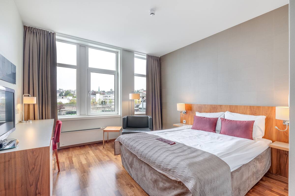 A hotel room with a large bed, a desk with a chair, a TV, and a window with curtains.