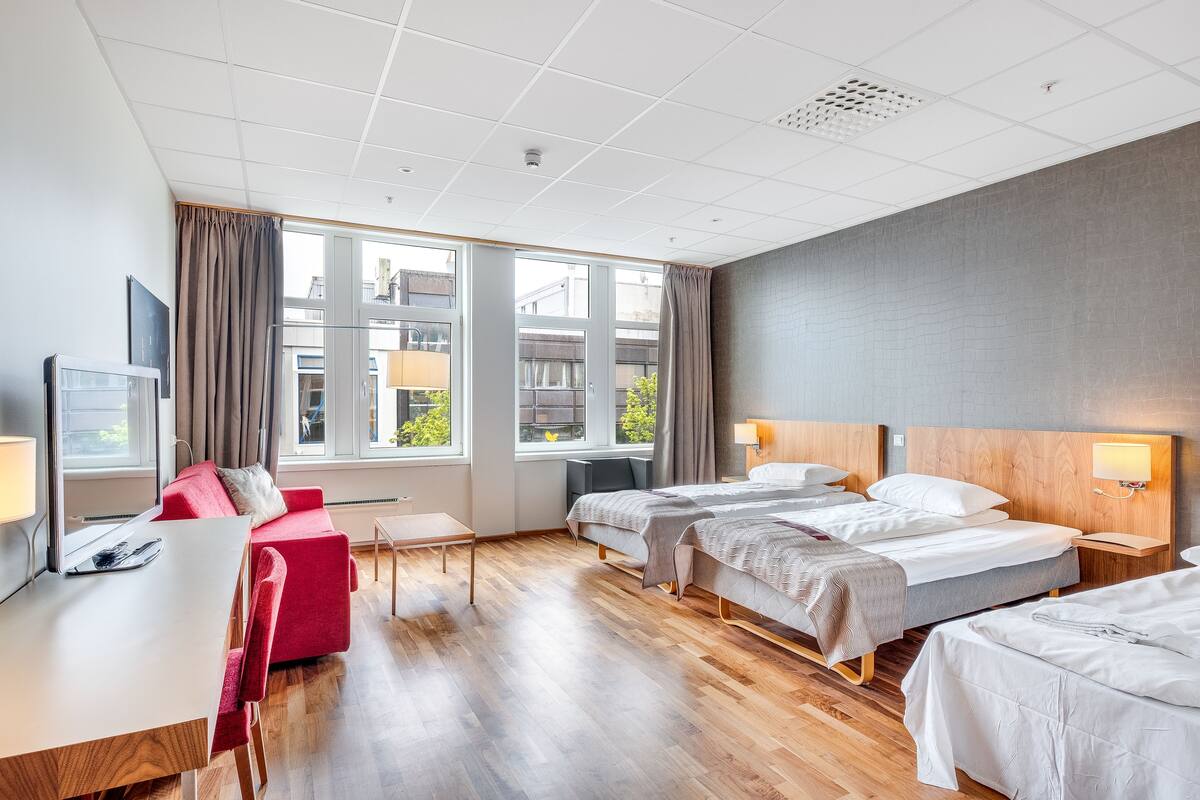 A hotel room with two beds, a desk, a red sofa, and large windows.
