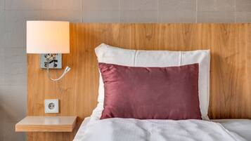 A neatly made bed with white and maroon pillows, a bedside table with a lamp, and a power outlet on the wall.