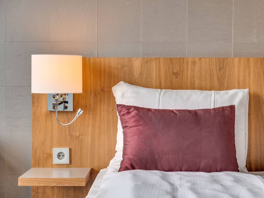 A neatly made bed with white and maroon pillows, a bedside table with a lamp, and a power outlet on the wall.