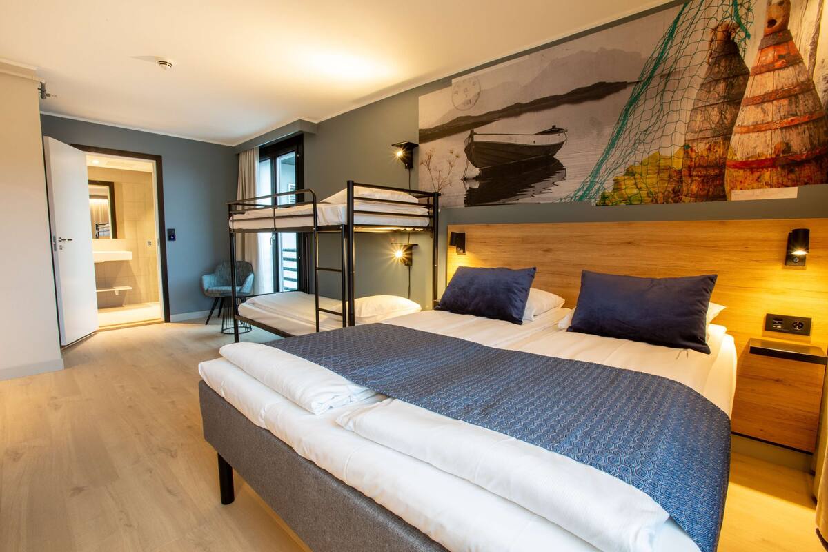 Une chambre d’hôtel moderne avec un lit superposé, un grand lit recouvert d’une couverture à motifs, une tête de lit en bois, une table de chevet et un tableau mural représentant des bateaux.