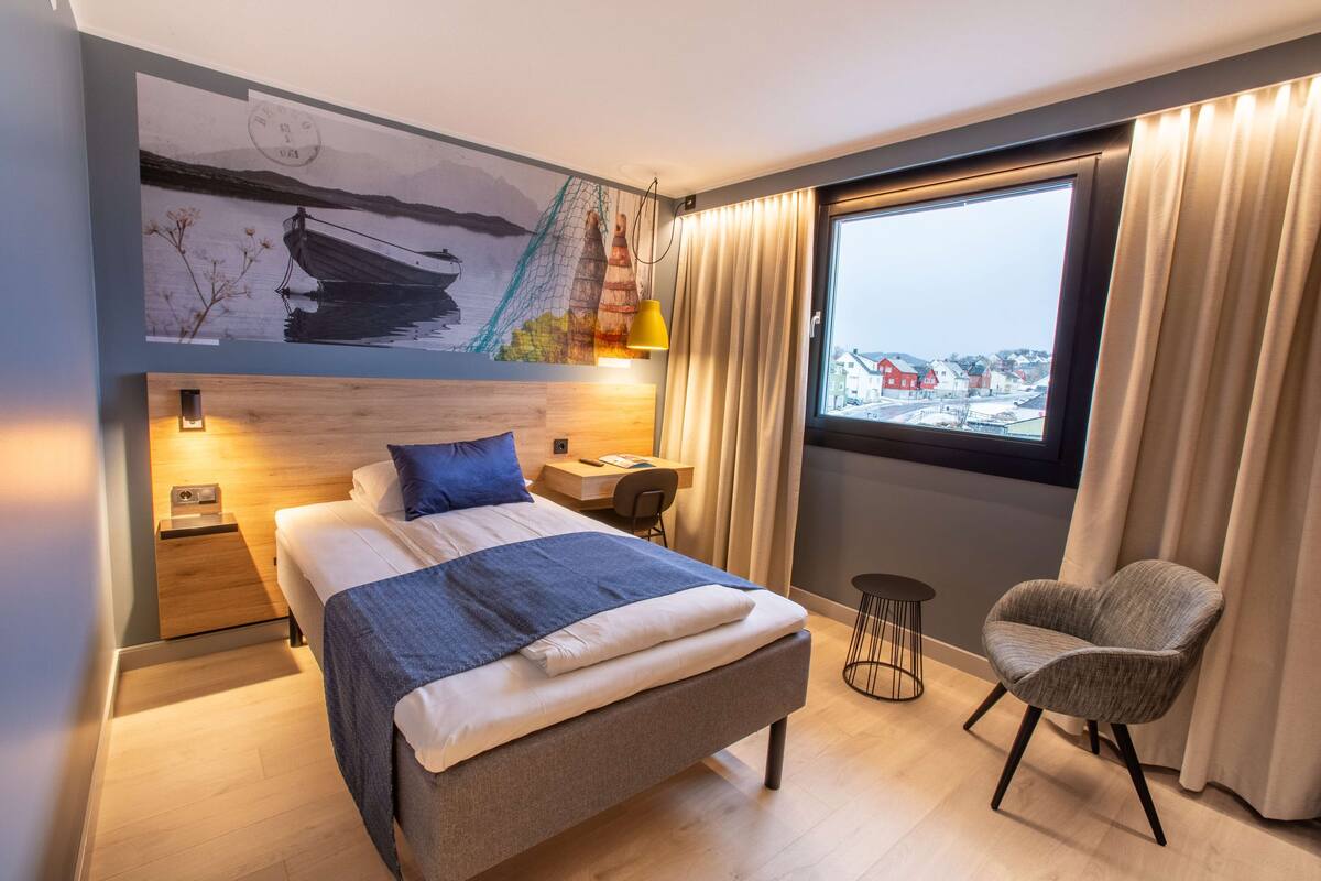 Une chambre d’hôtel avec un lit, une chaise, un bureau et une grande fenêtre avec des rideaux.