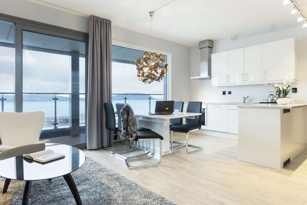 A modern kitchen with white cabinets, a dining area with a glass table, and a view through large windows.